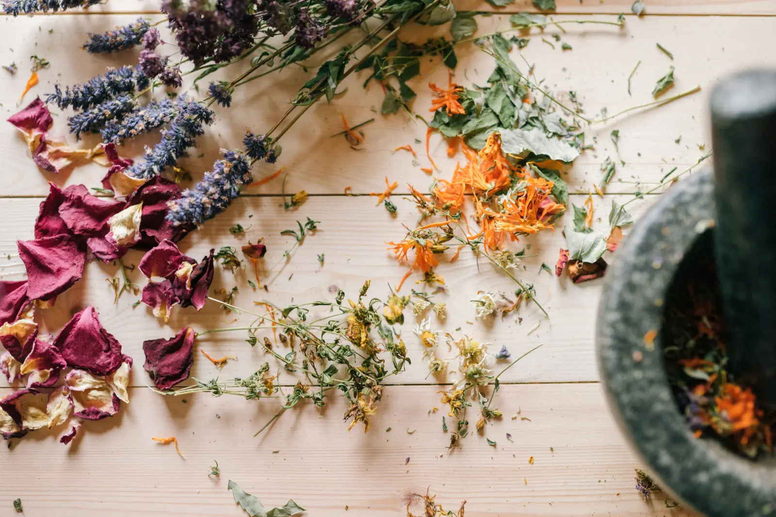 Getrocknete Kräuter, Lavendel, Rosenblütenblätter und Ringelblumen mit einem Mörser auf einem hellen Holztisch